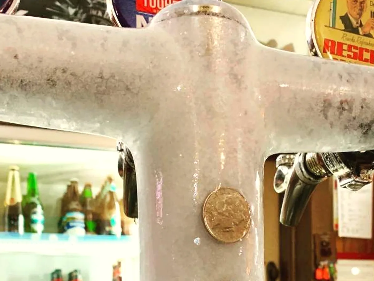 A frosted beer tap on the bar with coins stuck to the ice on it.