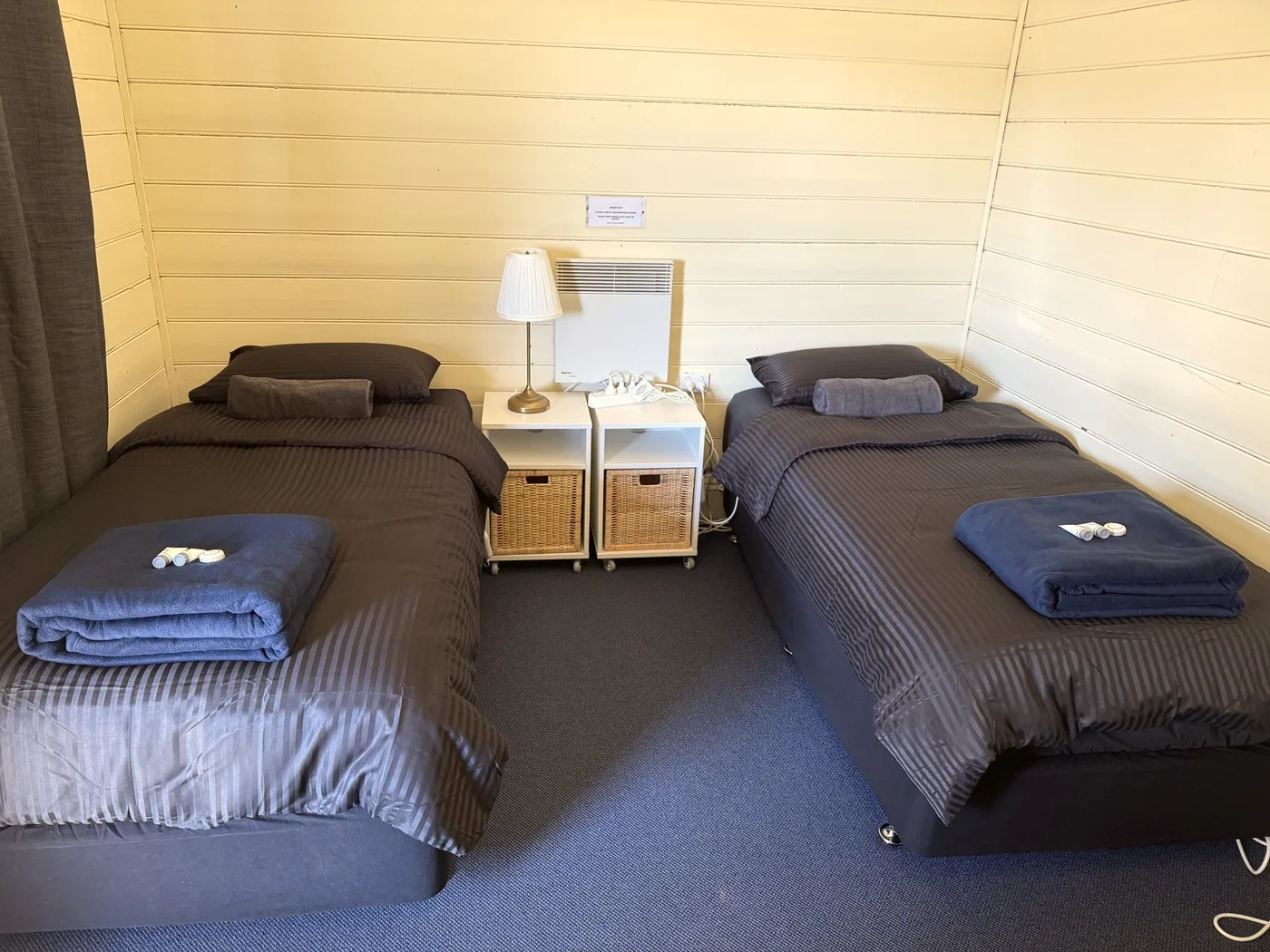 Two single beds in a cottage room with a curtain drawn across the window and towels folded on each bed.