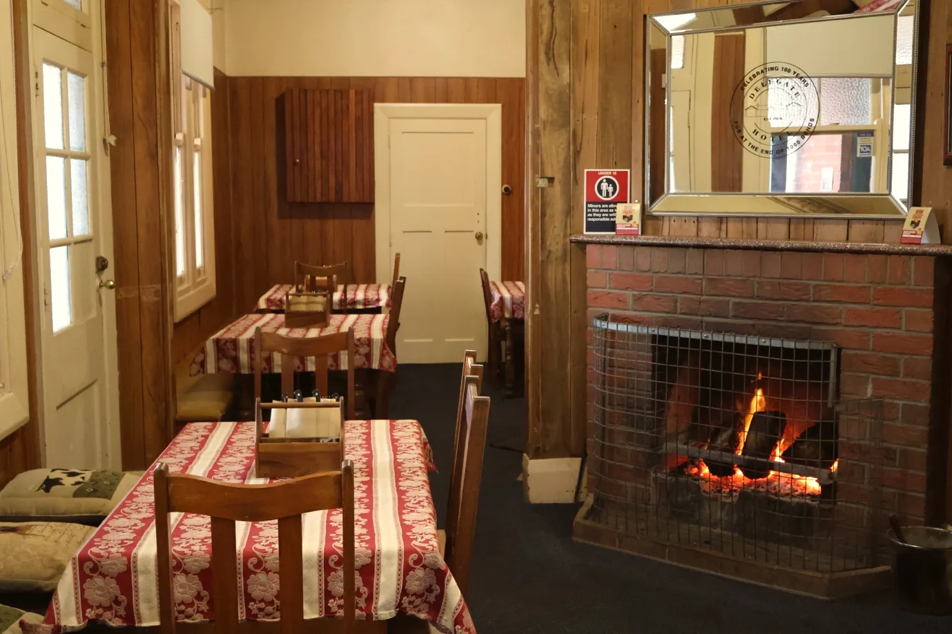 A brick fireplace with the fire lit, and the dining room beside it: tables under red and white patterned cloths, timber chairs and a mirror above the mantel carrying the hotel's centenary decal.