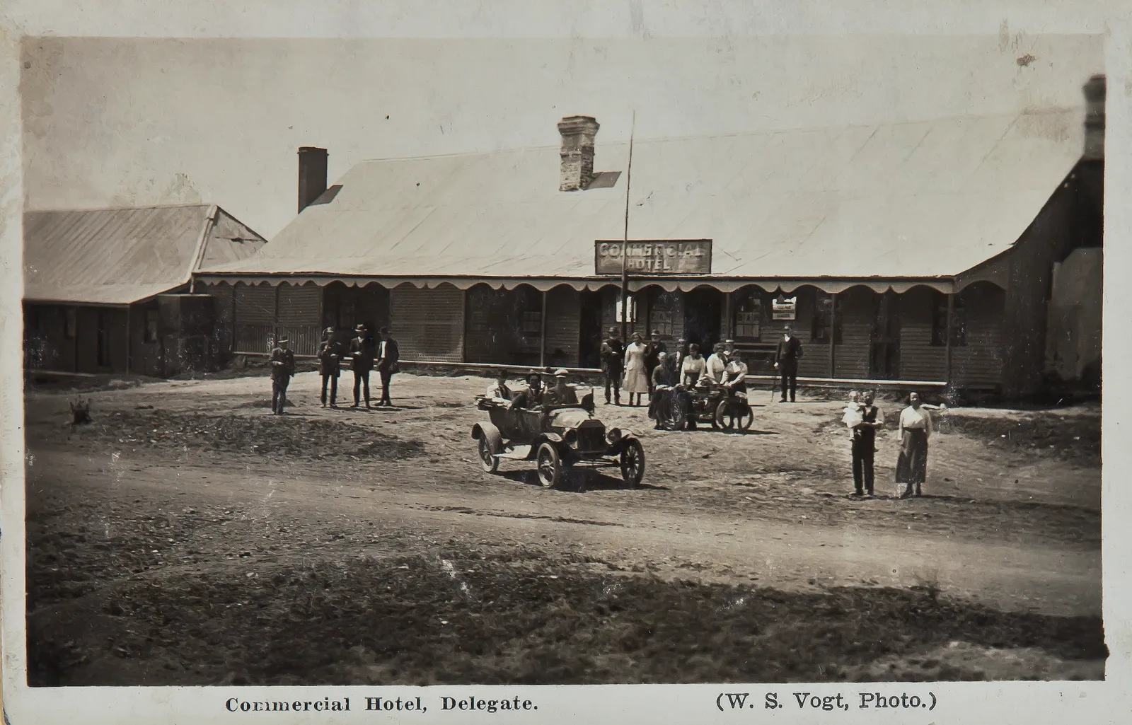 A period postcard of the Commercial Hotel, Delegate: a long single storey building with a scalloped veranda and a corrugated iron roof, with about twenty people, an open-top motorcar and a motorcycle and sidecar on the dirt road outside.