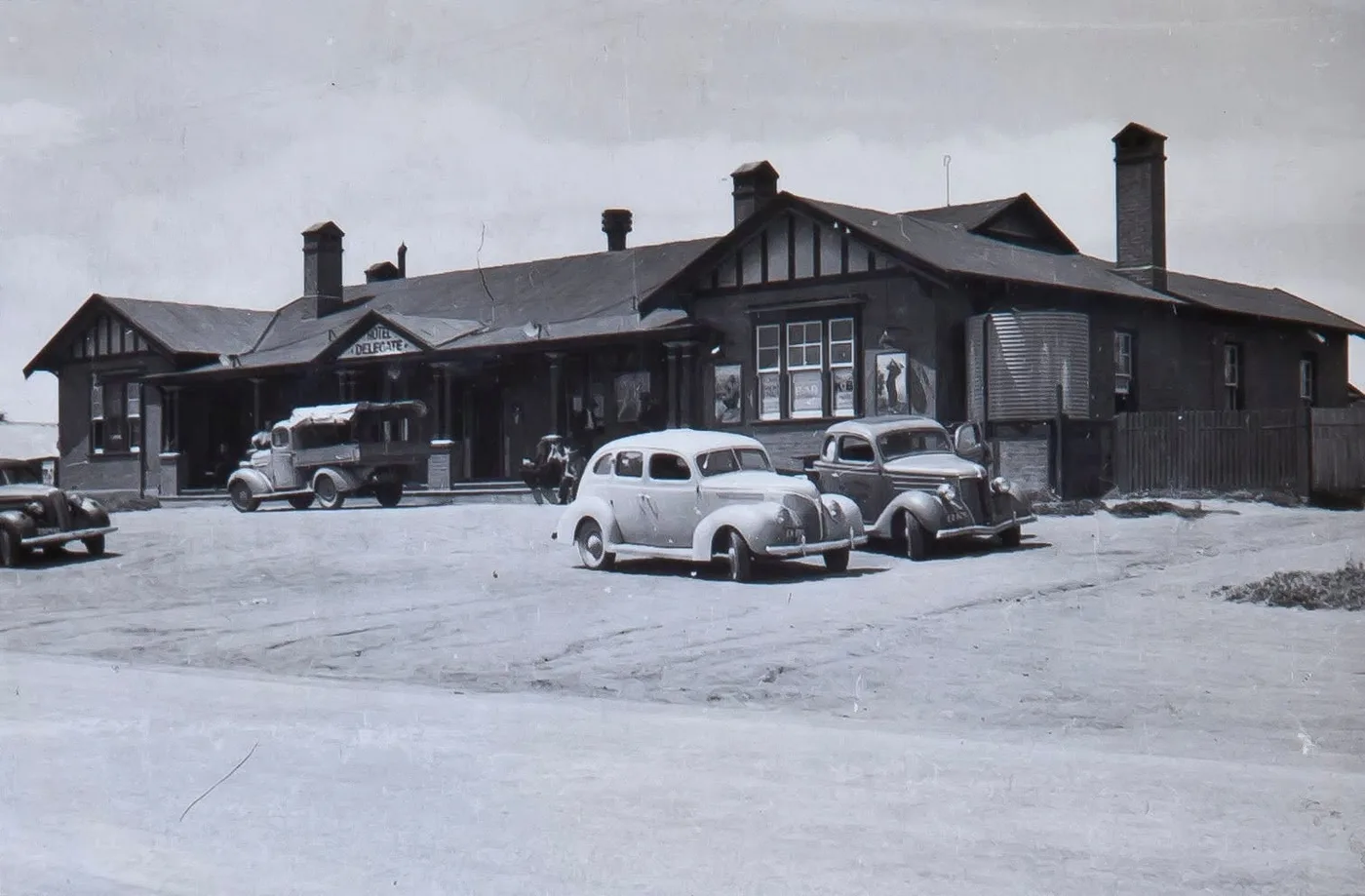 The rebuilt Delegate Hotel photographed in black and white: a long building with gabled, half-timbered ends and brick chimneys, with three period cars and a loaded flatbed truck parked on the gravel forecourt.