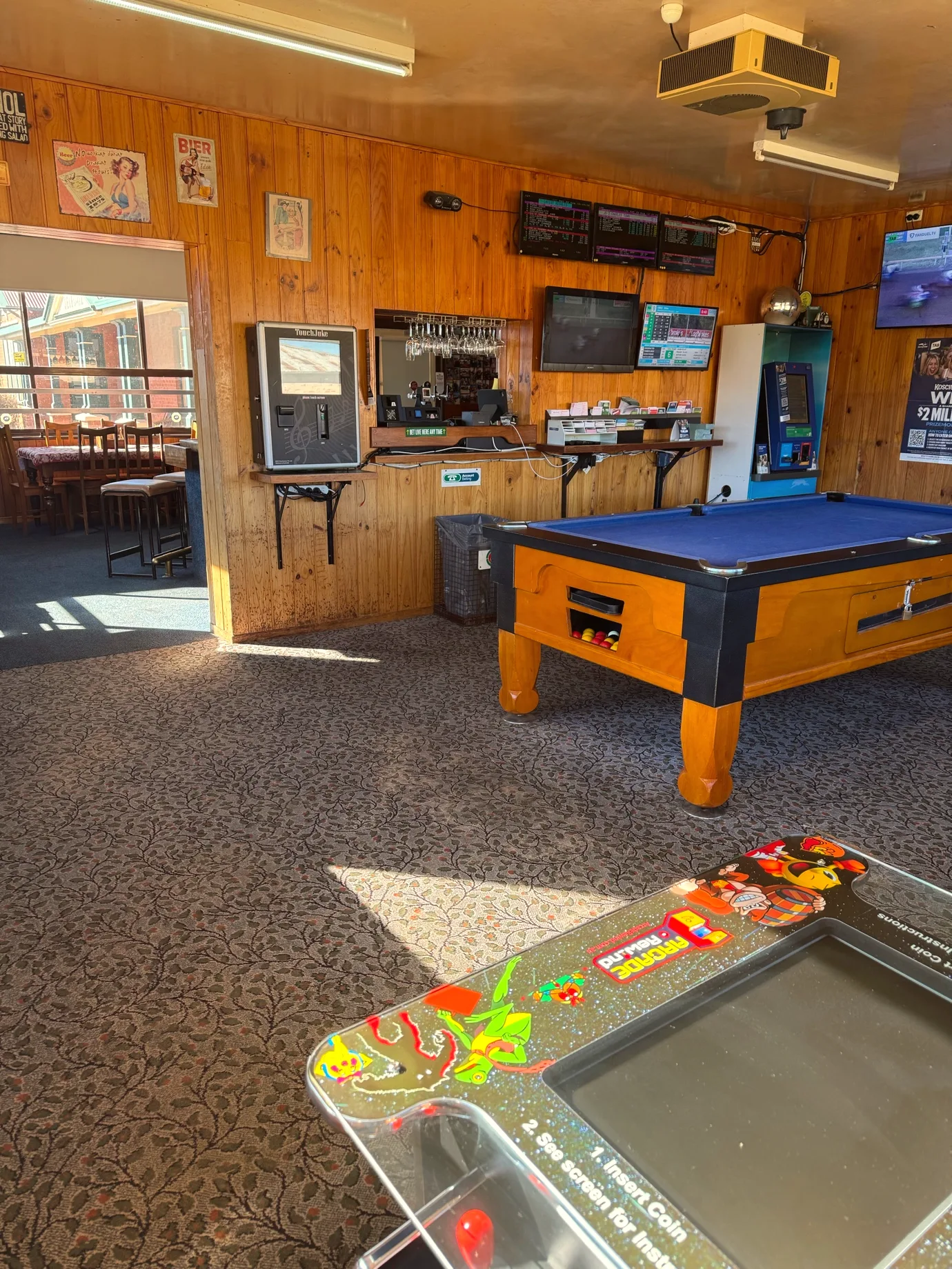 The pool room at the Delegate Hotel: a pool table with blue cloth, timber panelled walls, screens above the bar and a tabletop arcade cabinet in the foreground.