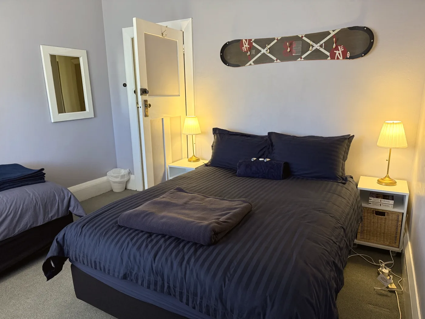 A hotel room with a queen bed made up in navy, a single bed alongside, lamps either side and a snowboard mounted on the wall.